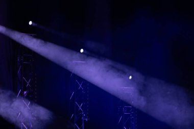 Mor sahne ışıkları sisli sisi delip geçer, karanlık bir tiyatro ortamında metal kirişleri vurgular. Güçlü atmosfer ve aydınlatma efektleri.