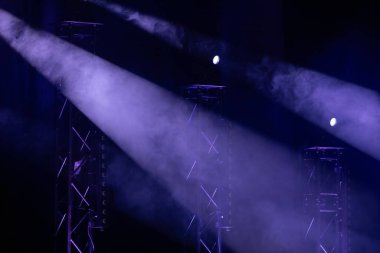 Mor sahne ışıkları sisli sisi delip geçer, karanlık bir tiyatro ortamında metal kirişleri vurgular. Güçlü atmosfer ve aydınlatma efektleri.