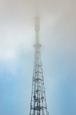 Yüksek bir iletişim kulesi yoğun sislerin içinden sislerin kısmen gizleyip gizemli ve atmosferik bir sahne oluşturduğu soluk mavi gökyüzüne doğru yükselir. Kopyalama alanı içerir.