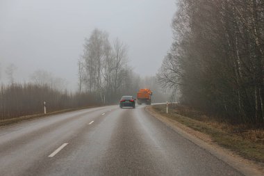 Siyah bir araba ve turuncu bir kamyon nemli, sislerle kaplı orman yolu boyunca seyahat ediyor. Görüş mesafesi sınırlı ve kışın soluk renkleri var. 03.26.2025 Limbazi, Letonya