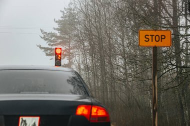 Siyah bir araba, çıplak ağaçlarla kaplı sisli bir kırsal yolda sarı bir dur işaretinin yanında kırmızı bir trafik lambasının önünde bekliyor. Huysuz ve atmosferik. Orman çevrili..