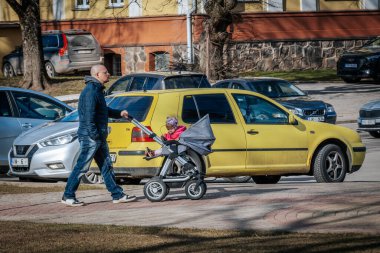  Kapüşonlu ve kotlu bir adam, güneşli bir günde sarı bir otoparkta, bebek arabasıyla bir bebek arabasını kenara itiyor. 03.27.2025 Valmiera, Letonya