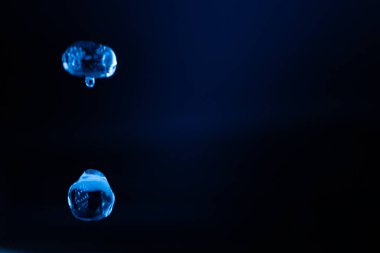 İki şeffaf mavi sıvı damlacık karanlık, minimum bir ortamda asılı, yansımalarla yavaşça parlayan ve geleceksel, ruhani bir hava yaratıyor. Kopyalama alanı içerir.