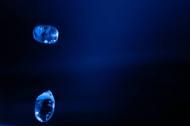 İki şeffaf mavi sıvı damlacık karanlık, minimum bir ortamda asılı, yansımalarla yavaşça parlayan ve geleceksel, ruhani bir hava yaratıyor. Kopyalama alanı içerir.