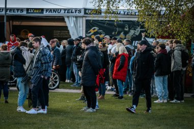 Bir grup insan büyük bir LED ekranda spor maçı izlemek için dışarıda toplandı. Kırmızı elbiseli taraftarlar tezahürat yapıyordu. Hokey hayranları canlı yayın izliyor. 10.05.2025 Valmiera, Letonya