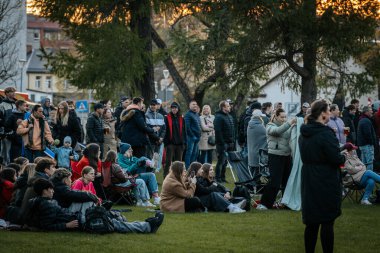 Çoğu battaniye ve sırt çantalarıyla çimlerin üzerinde oturan bir grup insan, serin havada bir açık hava etkinliğini dikkatle izliyor. 10.05.2025 Valmiera, Letonya