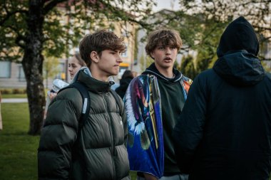 Çoğu battaniye ve sırt çantalarıyla çimlerin üzerinde oturan bir grup insan, serin havada bir açık hava etkinliğini dikkatle izliyor. 10.05.2025 Valmiera, Letonya