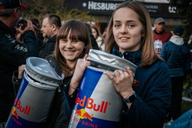 İki genç kadın, Red Bull markalı soğutucuları kalabalık bir açık hava ortamında tutarken gülümsüyor, muhtemelen bir promosyon veya spor etkinliği sırasında. 10.05.2025 Valmiera, Letonya