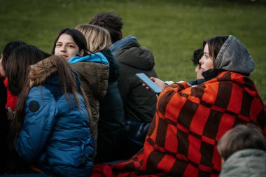 Çoğu battaniye ve sırt çantalarıyla çimlerin üzerinde oturan bir grup insan, serin havada bir açık hava etkinliğini dikkatle izliyor. 10.05.2025 Valmiera, Letonya
