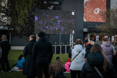 Bir grup insan büyük bir LED ekranda spor maçı izlemek için dışarıda toplandı. Kırmızı elbiseli taraftarlar tezahürat yapıyordu. Hokey hayranları canlı yayın izliyor. 10.05.2025 Valmiera, Letonya