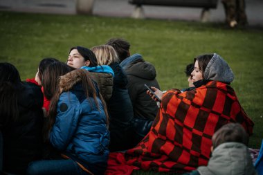Çoğu battaniye ve sırt çantalarıyla çimlerin üzerinde oturan bir grup insan, serin havada bir açık hava etkinliğini dikkatle izliyor. 10.05.2025 Valmiera, Letonya