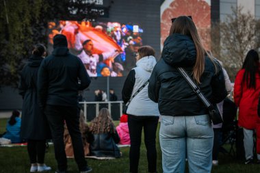 Bir grup insan büyük bir LED ekranda spor maçı izlemek için dışarıda toplandı. Kırmızı elbiseli taraftarlar tezahürat yapıyordu. Hokey hayranları canlı yayın izliyor. 10.05.2025 Valmiera, Letonya