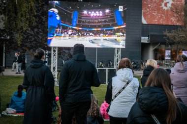 Bir grup insan büyük bir LED ekranda spor maçı izlemek için dışarıda toplandı. Kırmızı elbiseli taraftarlar tezahürat yapıyordu. Hokey hayranları canlı yayın izliyor. 10.05.2025 Valmiera, Letonya