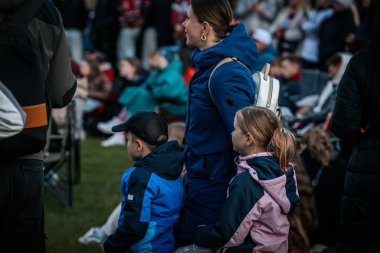 Bir grup insan büyük bir LED ekranda spor maçı izlemek için dışarıda toplandı. Kırmızı elbiseli taraftarlar tezahürat yapıyordu. Hokey hayranları canlı yayın izliyor. 10.05.2025 Valmiera, Letonya