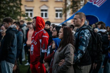 Bir grup insan büyük bir LED ekranda spor maçı izlemek için dışarıda toplandı. Kırmızı elbiseli taraftarlar tezahürat yapıyordu. Hokey hayranları canlı yayın izliyor. 10.05.2025 Valmiera, Letonya