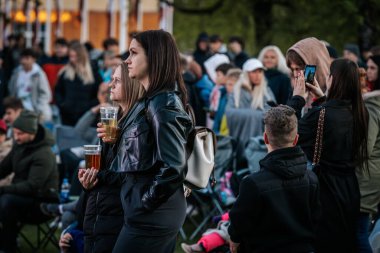 Bir grup insan büyük bir LED ekranda spor maçı izlemek için dışarıda toplandı. Kırmızı elbiseli taraftarlar tezahürat yapıyordu. Hokey hayranları canlı yayın izliyor. 10.05.2025 Valmiera, Letonya