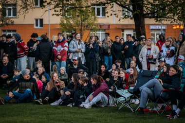 Bir grup insan büyük bir LED ekranda spor maçı izlemek için dışarıda toplandı. Kırmızı elbiseli taraftarlar tezahürat yapıyordu. Hokey hayranları canlı yayın izliyor. 10.05.2025 Valmiera, Letonya