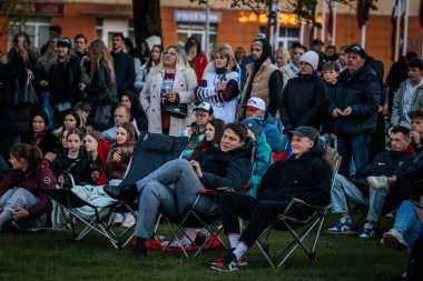Bir grup insan büyük bir LED ekranda spor maçı izlemek için dışarıda toplandı. Kırmızı elbiseli taraftarlar tezahürat yapıyordu. Hokey hayranları canlı yayın izliyor. 10.05.2025 Valmiera, Letonya