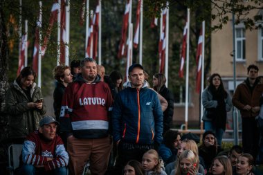 Bir grup insan büyük bir LED ekranda spor maçı izlemek için dışarıda toplandı. Kırmızı elbiseli taraftarlar tezahürat yapıyordu. Hokey hayranları canlı yayın izliyor. 10.05.2025 Valmiera, Letonya