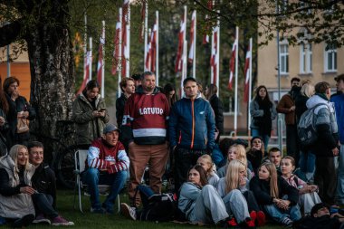 Bir grup insan büyük bir LED ekranda spor maçı izlemek için dışarıda toplandı. Kırmızı elbiseli taraftarlar tezahürat yapıyordu. Hokey hayranları canlı yayın izliyor. 10.05.2025 Valmiera, Letonya