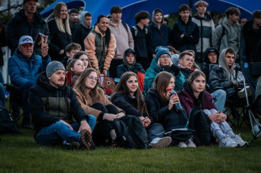 Çoğu battaniye ve sırt çantalarıyla çimlerin üzerinde oturan bir grup insan, serin havada bir açık hava etkinliğini dikkatle izliyor. 10.05.2025 Valmiera, Letonya