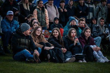 Çoğu battaniye ve sırt çantalarıyla çimlerin üzerinde oturan bir grup insan, serin havada bir açık hava etkinliğini dikkatle izliyor. 10.05.2025 Valmiera, Letonya