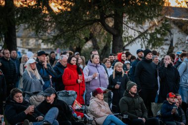 Çoğu battaniye ve sırt çantalarıyla çimlerin üzerinde oturan bir grup insan, serin havada bir açık hava etkinliğini dikkatle izliyor. 10.05.2025 Valmiera, Letonya