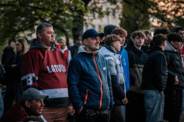 Bir grup insan büyük bir LED ekranda spor maçı izlemek için dışarıda toplandı. Kırmızı elbiseli taraftarlar tezahürat yapıyordu. Hokey hayranları canlı yayın izliyor. 10.05.2025 Valmiera, Letonya