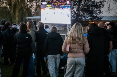 Bir grup insan büyük bir LED ekranda spor maçı izlemek için dışarıda toplandı. Kırmızı elbiseli taraftarlar tezahürat yapıyordu. Hokey hayranları canlı yayın izliyor. 10.05.2025 Valmiera, Letonya