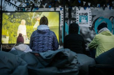 Sıcak ceketli ve şapkalı insanlar, Kino Pedlis festivali sırasında açık ekrana yansıtılan ve rahat armut çuvalları ve bayrak ile çevrili bir film izliyorlar. 10.05.2025 Valmiera, Letonya