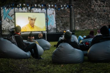 Sıcak ceketli ve şapkalı insanlar, Kino Pedlis festivali sırasında açık ekrana yansıtılan ve rahat armut çuvalları ve bayrak ile çevrili bir film izliyorlar. 10.05.2025 Valmiera, Letonya