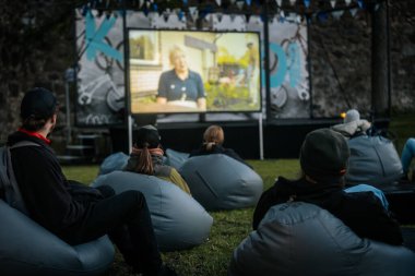 Sıcak ceketli ve şapkalı insanlar, Kino Pedlis festivali sırasında açık ekrana yansıtılan ve rahat armut çuvalları ve bayrak ile çevrili bir film izliyorlar. 10.05.2025 Valmiera, Letonya