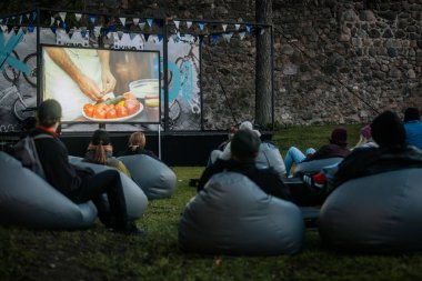 Sıcak ceketli ve şapkalı insanlar, Kino Pedlis festivali sırasında açık ekrana yansıtılan ve rahat armut çuvalları ve bayrak ile çevrili bir film izliyorlar. 10.05.2025 Valmiera, Letonya