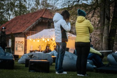 Sıcak ceketli ve şapkalı insanlar, Kino Pedlis festivali sırasında açık ekrana yansıtılan ve rahat armut çuvalları ve bayrak ile çevrili bir film izliyorlar. 10.05.2025 Valmiera, Letonya