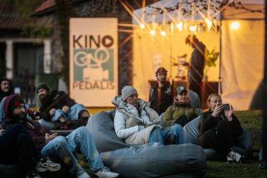 Sıcak ceketli ve şapkalı insanlar, Kino Pedlis festivali sırasında açık ekrana yansıtılan ve rahat armut çuvalları ve bayrak ile çevrili bir film izliyorlar. 10.05.2025 Valmiera, Letonya