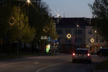 Sakin bir şehir caddesi, alacakaranlıkta yıldız patlaması desenli sokak lambalarıyla parıldıyor, yeşil ağaçlarla kaplı ve solgun bir gökyüzünün altında binalarla çevrili. 10.05.2025 Valmiera, Letonya