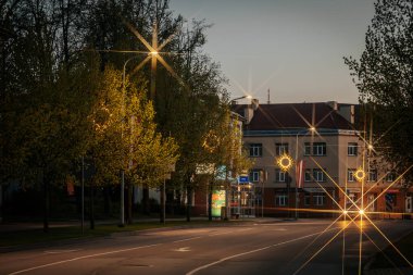 Sakin bir şehir caddesi, alacakaranlıkta yıldız patlaması desenli sokak lambalarıyla parıldıyor, yeşil ağaçlarla kaplı ve solgun bir gökyüzünün altında binalarla çevrili. 10.05.2025 Valmiera, Letonya