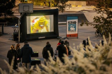 Bir grup insan büyük bir LED ekranda spor maçı izlemek için dışarıda toplandı. Kırmızı elbiseli taraftarlar tezahürat yapıyordu. Hokey hayranları canlı yayın izliyor. 10.05.2025 Valmiera, Letonya