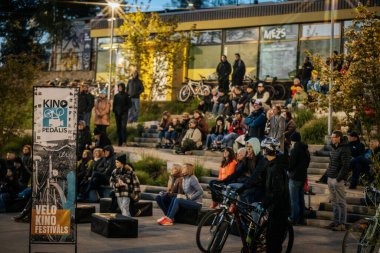 İnsanlar Kino Pedalis festivali sırasında ön planda tanıtım pankartıyla merdivenlerde oturup açık hava film gösterimi izliyor. 10.05.2025 Valmiera, Letonya