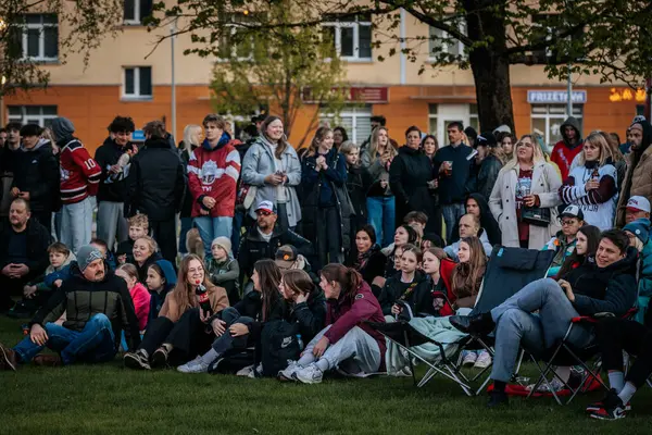 Bir grup insan büyük bir LED ekranda spor maçı izlemek için dışarıda toplandı. Kırmızı elbiseli taraftarlar tezahürat yapıyordu. Hokey hayranları canlı yayın izliyor. 10.05.2025 Valmiera, Letonya