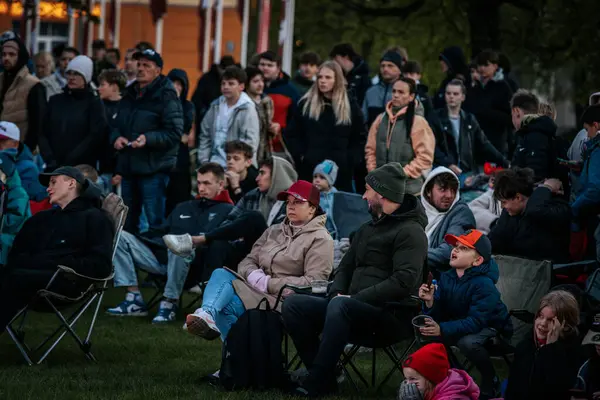 Bir grup insan büyük bir LED ekranda spor maçı izlemek için dışarıda toplandı. Kırmızı elbiseli taraftarlar tezahürat yapıyordu. Hokey hayranları canlı yayın izliyor. 10.05.2025 Valmiera, Letonya