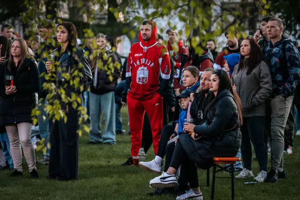 Bir grup insan büyük bir LED ekranda spor maçı izlemek için dışarıda toplandı. Kırmızı elbiseli taraftarlar tezahürat yapıyordu. Hokey hayranları canlı yayın izliyor. 10.05.2025 Valmiera, Letonya