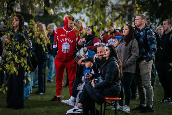 Bir grup insan büyük bir LED ekranda spor maçı izlemek için dışarıda toplandı. Kırmızı elbiseli taraftarlar tezahürat yapıyordu. Hokey hayranları canlı yayın izliyor. 10.05.2025 Valmiera, Letonya