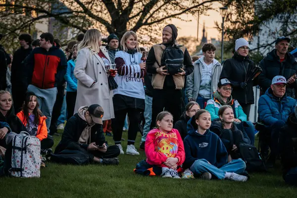Çoğu battaniye ve sırt çantalarıyla çimlerin üzerinde oturan bir grup insan, serin havada bir açık hava etkinliğini dikkatle izliyor. 10.05.2025 Valmiera, Letonya