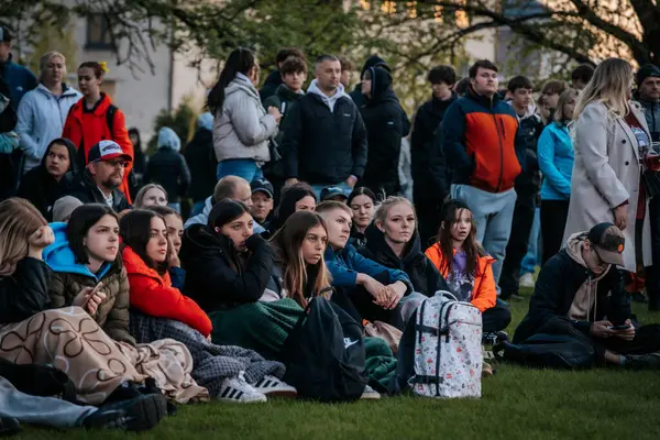 Çoğu battaniye ve sırt çantalarıyla çimlerin üzerinde oturan bir grup insan, serin havada bir açık hava etkinliğini dikkatle izliyor. 10.05.2025 Valmiera, Letonya