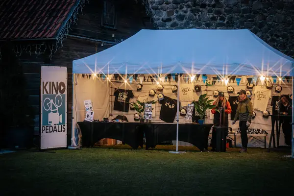 A vibrant street market in Limbazi features white tents, visitors browsing local goods, blooming flowers, and a purple balloon promoting tourism.
