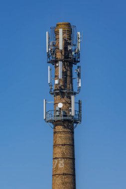 Birden fazla modern hücresel anten ve uydu antenleri ile donatılmış, parlak mavi gökyüzüne karşı yeniden tasarlanmış bir tuğla bacası. Cömert kopya alanı içerir.