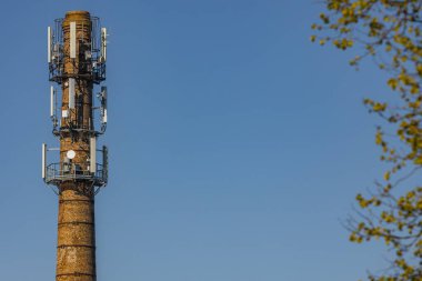 Modern hücre antenleri ve uydu antenleriyle donatılmış eski bir tuğla bacası. Önünde ağaç dalları olan açık mavi bir gökyüzüne karşı duruyor..