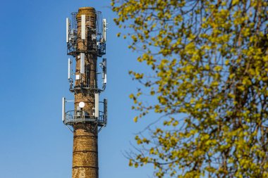 Modern hücre antenleri ve uydu antenleriyle donatılmış eski bir tuğla bacası. Önünde ağaç dalları olan açık mavi bir gökyüzüne karşı duruyor..