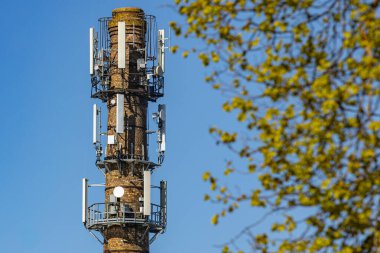 Modern hücre antenleri ve uydu antenleriyle donatılmış eski bir tuğla bacası. Önünde ağaç dalları olan açık mavi bir gökyüzüne karşı duruyor..